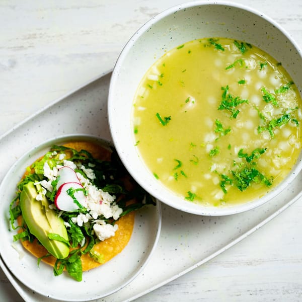 A Mexican tostada with avocado, lettuce, cheese, and radish, next to a bowl of pozole soup.