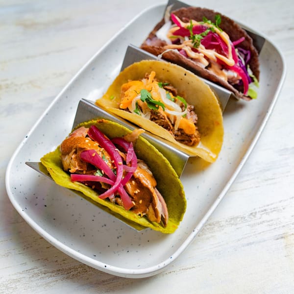 Three colorful tacos with various fillings and toppings, including pickled onions, on a white serving dish.