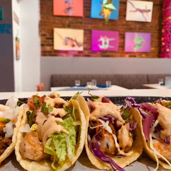 Close-up of four colorful tacos with various fillings and sauces, in a vibrant restaurant setting.