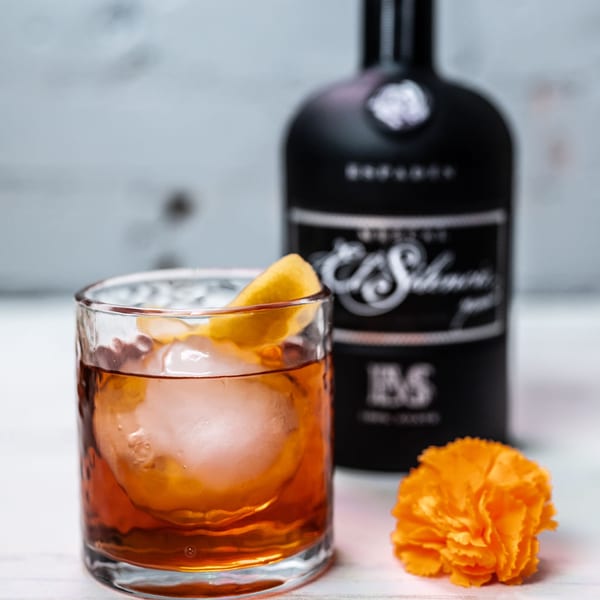 A cocktail with a large ice sphere and lemon peel, next to a black bottle and orange flower.