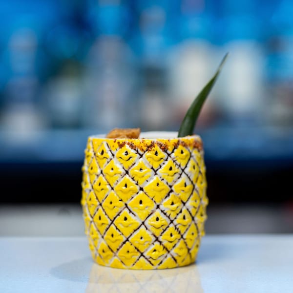 A pineapple-shaped cocktail glass with a drink, garnished with a leaf and a churro.