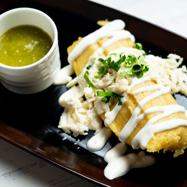 Mexican tamales topped with white sauce, cheese, and cilantro, with a side of green salsa.
